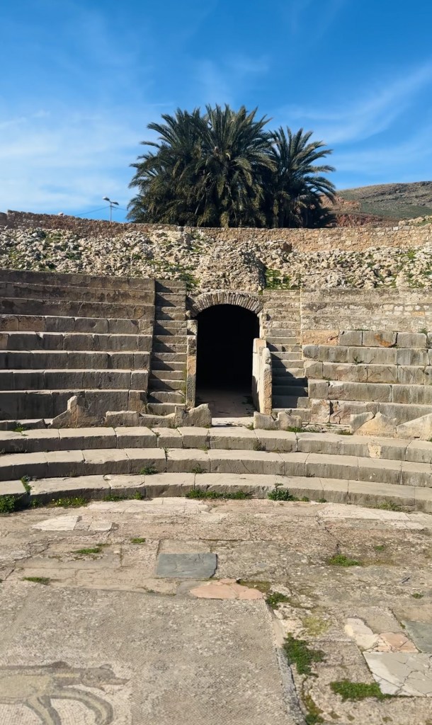 Ancient amphitheater entrance at Bulla Regia surrounded by stone steps and palm trees under a clear blue sky.