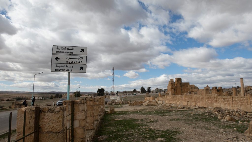 Road sign in Arabic and French pointing to 'Algerian Border,' 'El Ajrad,' and 'Centre Ville,' with ancient ruins and a cloudy sky in the background at Haïdra, Tunisia