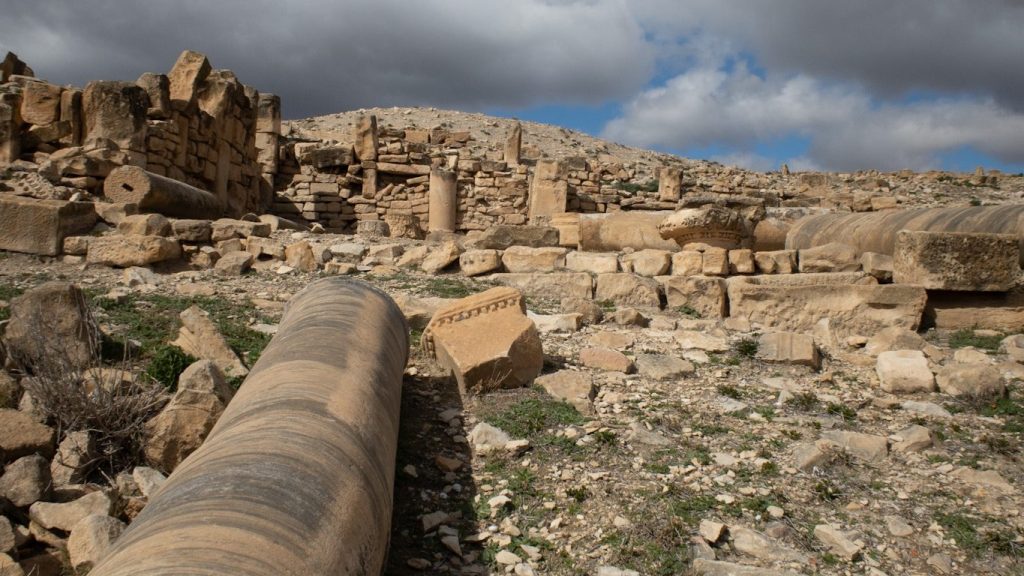 A large, fallen marble column with striped patterns lies in the foreground of extensive Roman ruins at Ammaedara in Haïdra, Tunisia