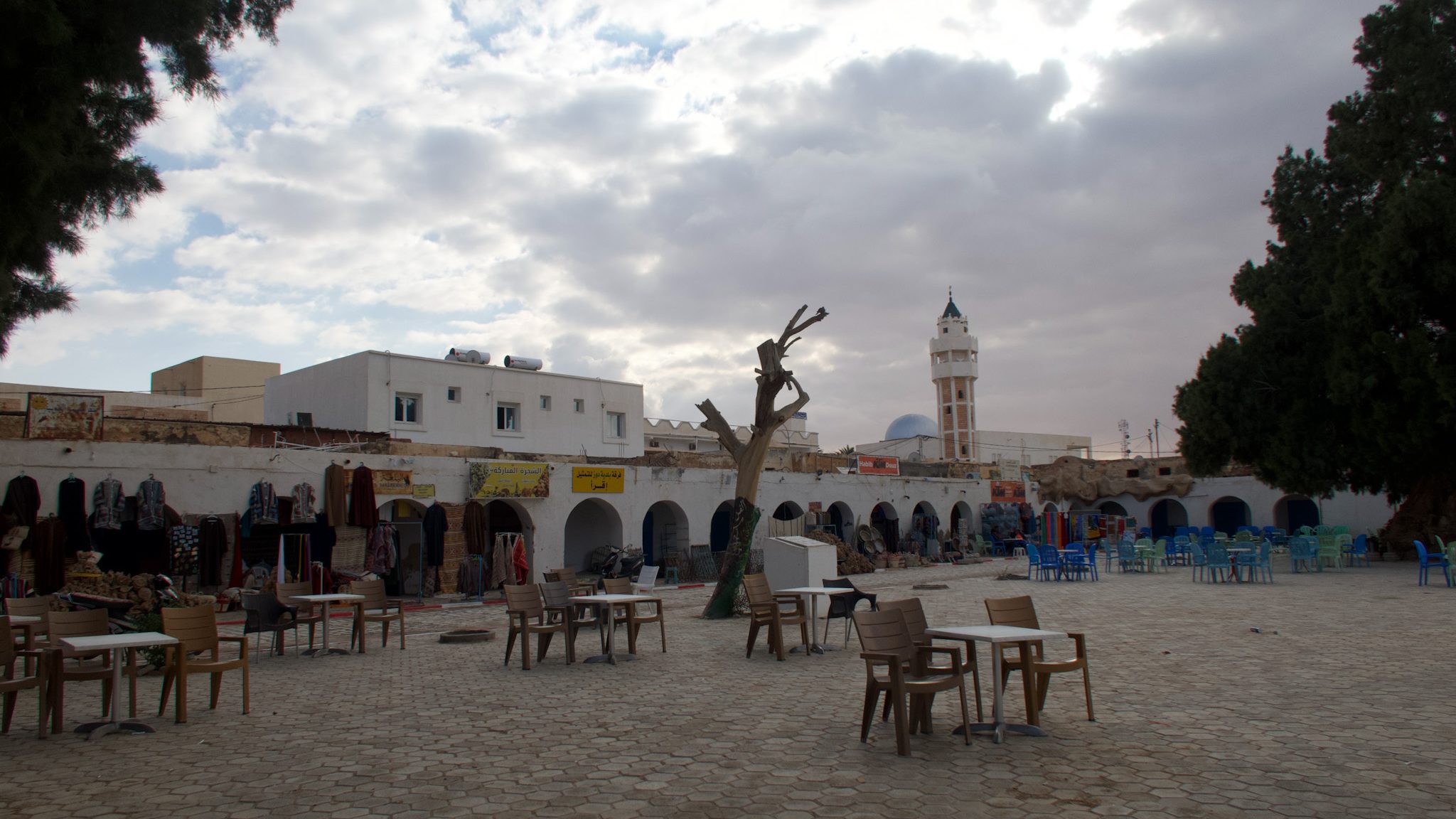 An open plaza in Douz, Tunisia featuring empty tables and chairs, surrounded by traditional market stalls and buildings with a mosque tower in the background under a cloudy sky.