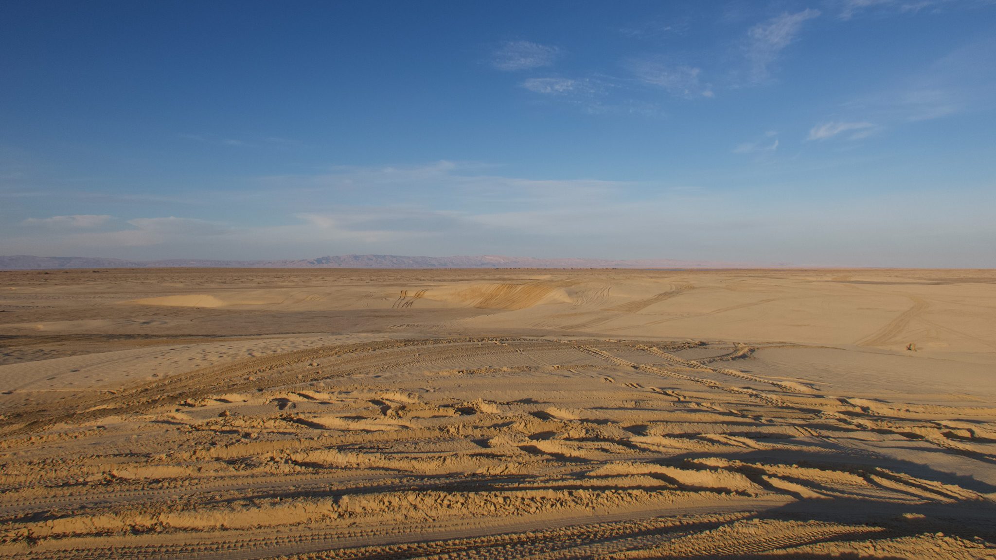 A vast desert landscape heading towards Ong Jemel in Tunisia, with clear blue skies and gentle curves of sand dunes, featuring tire tracks across the sandy terrain.