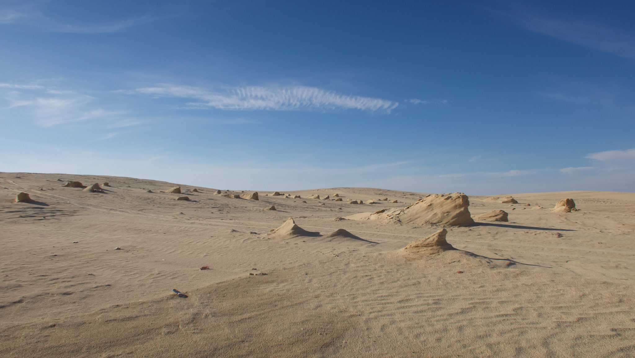 A vast desert landscape in the Tunisian Sahara featuring scattered rock formations under a blue sky with wispy clouds.