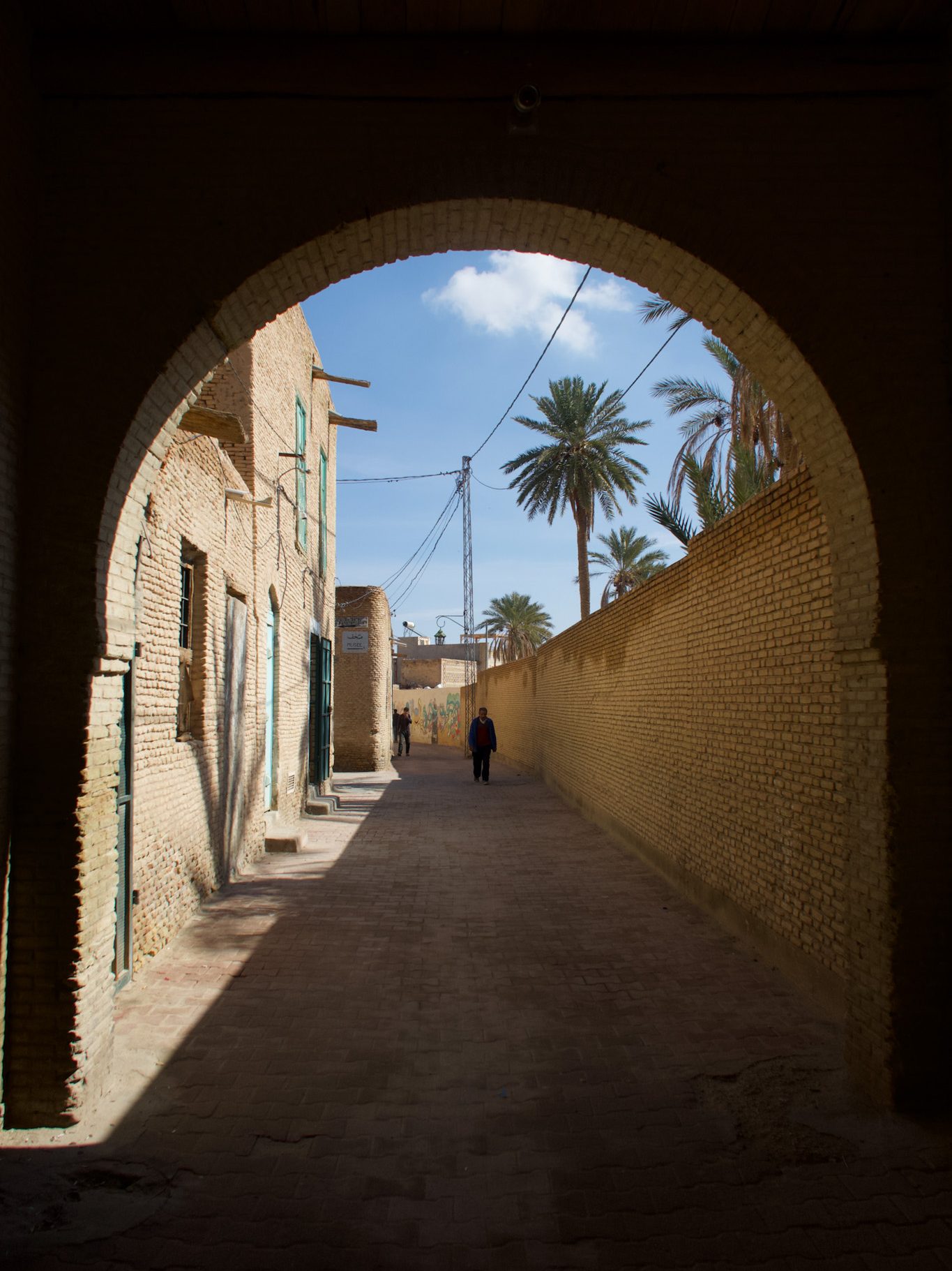 A narrow street view through an arched entrance in the Tozeur Medina, showcasing a sandy path lined with brick walls and palm trees in the background.