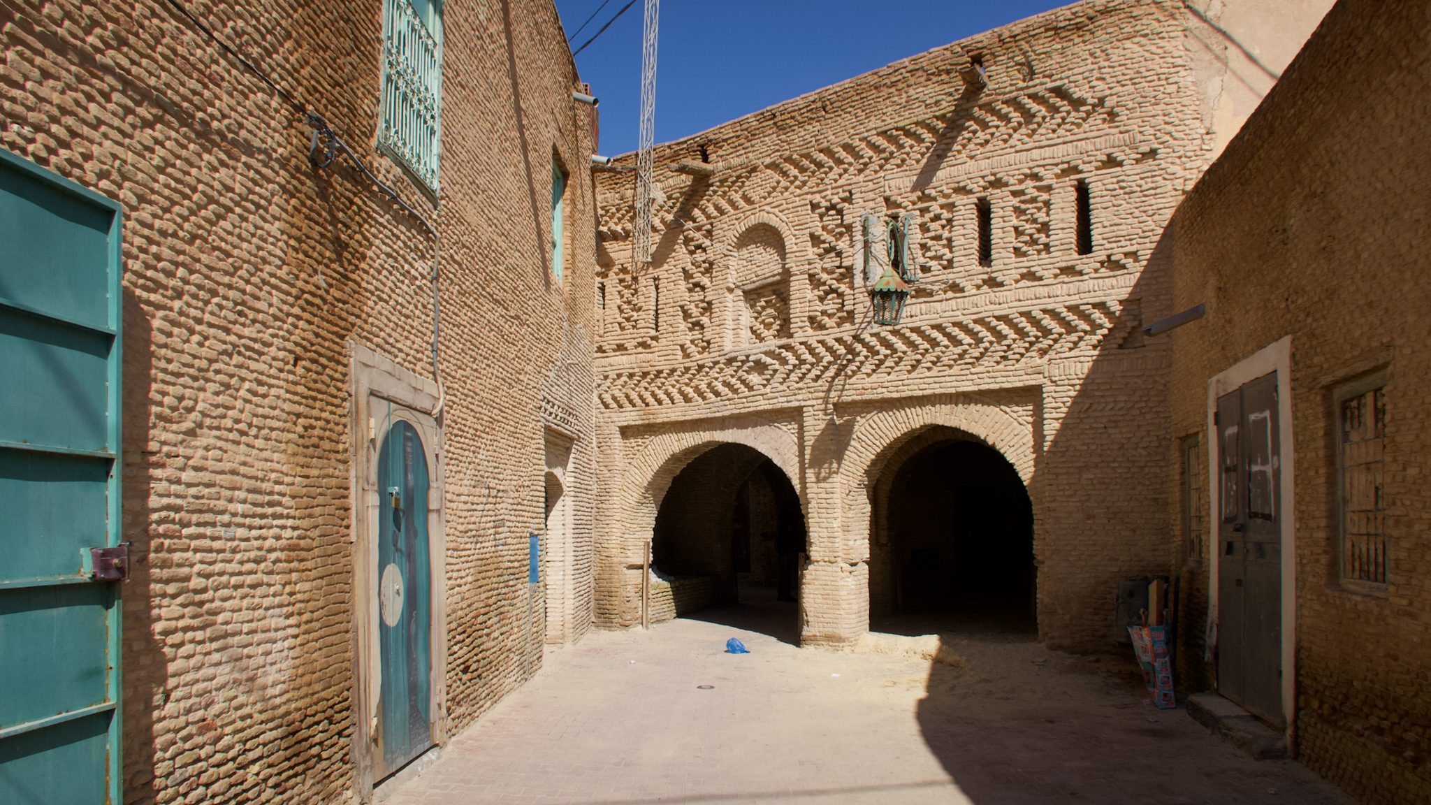 Narrow street in Tozeur, Tunisia with rustic brick buildings and decorative architecture, featuring arched doorways and a blue sky.