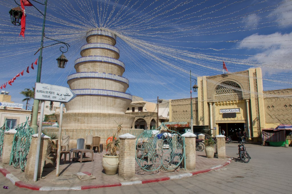 A large decorative fountain structure in the center of Tozeur's town square, with strings of lights overhead and surrounding buildings visible.
