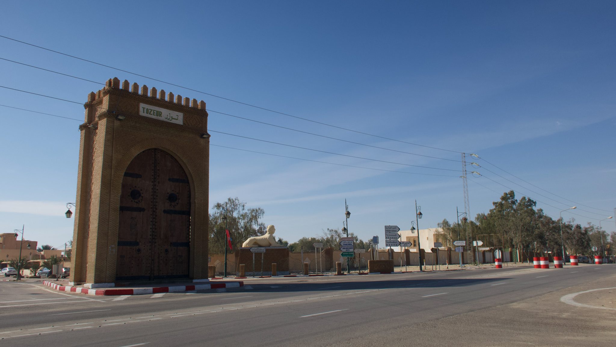 The entrance gate to Tozeur, Tunisia, featuring a large arched design and wooden doors, with road signs and a lion sculpture nearby.